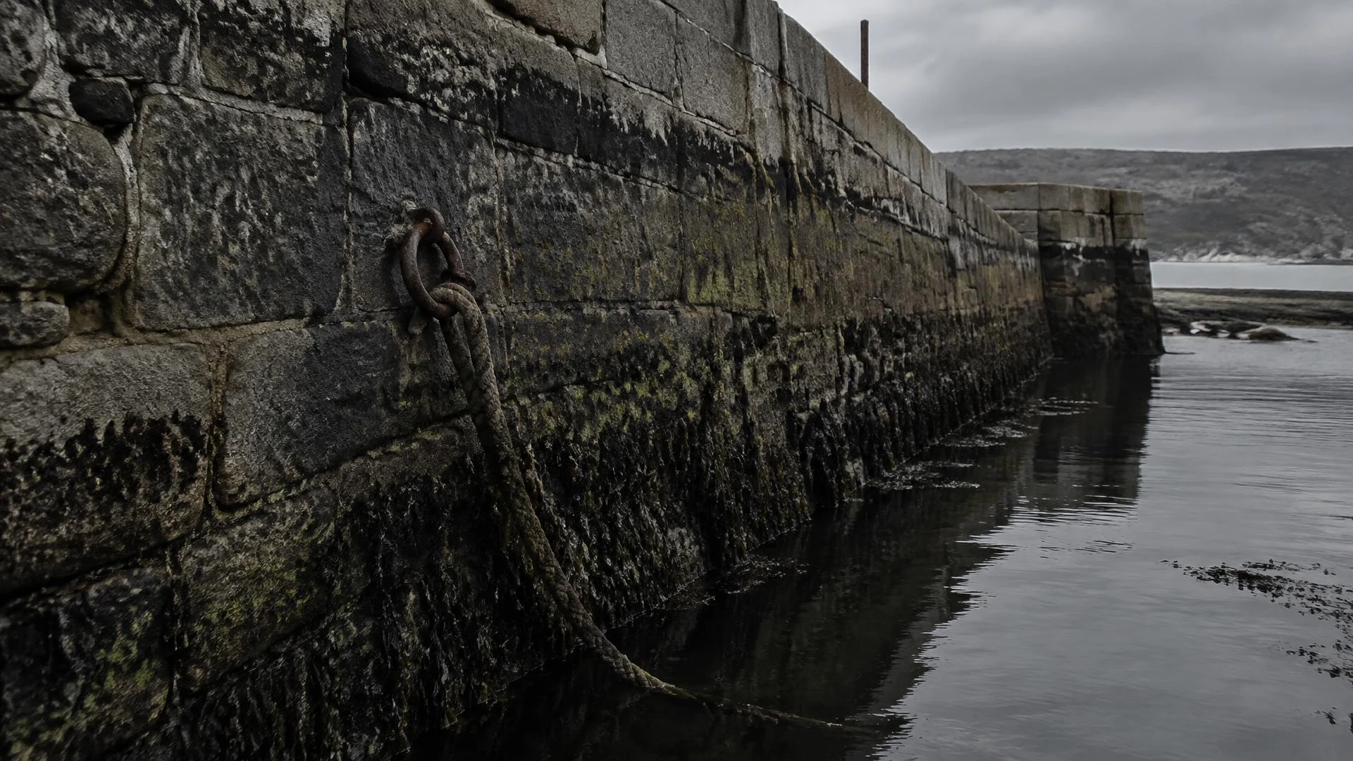 Illustration: a weathered stone harbour wall at low tide with a mooring ring and slack rope